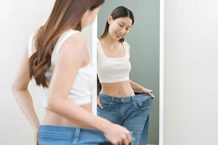 Woman smiling at her reflection in loose jeans, celebrating weight loss success from oral tirzepatide results.